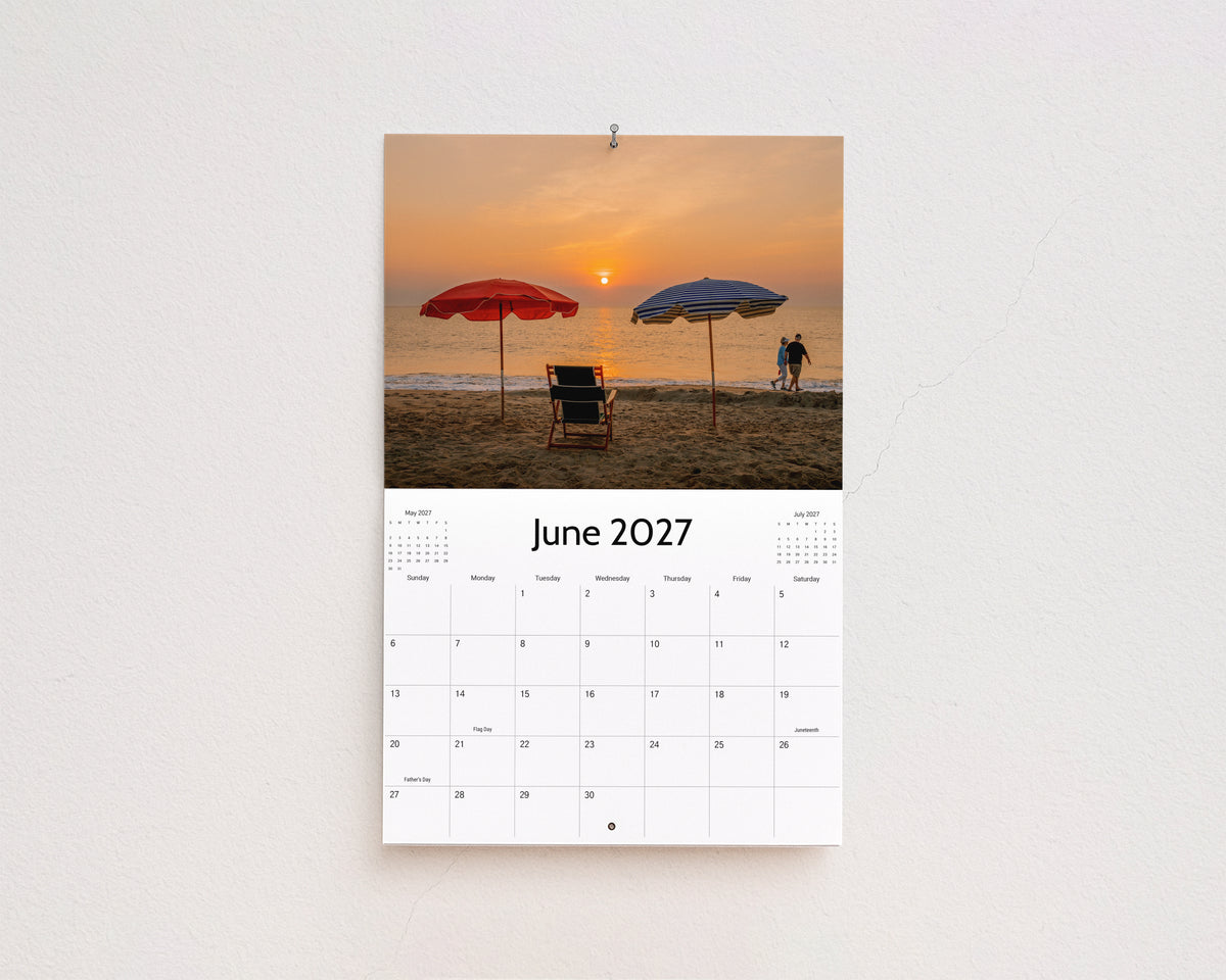 June 2027 page: red and blue beach umbrellas and a chair on the sand at sunrise
