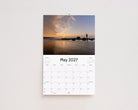 May 2027 page: a person standing on the rocks at sunrise with the sky reflected on wet sand