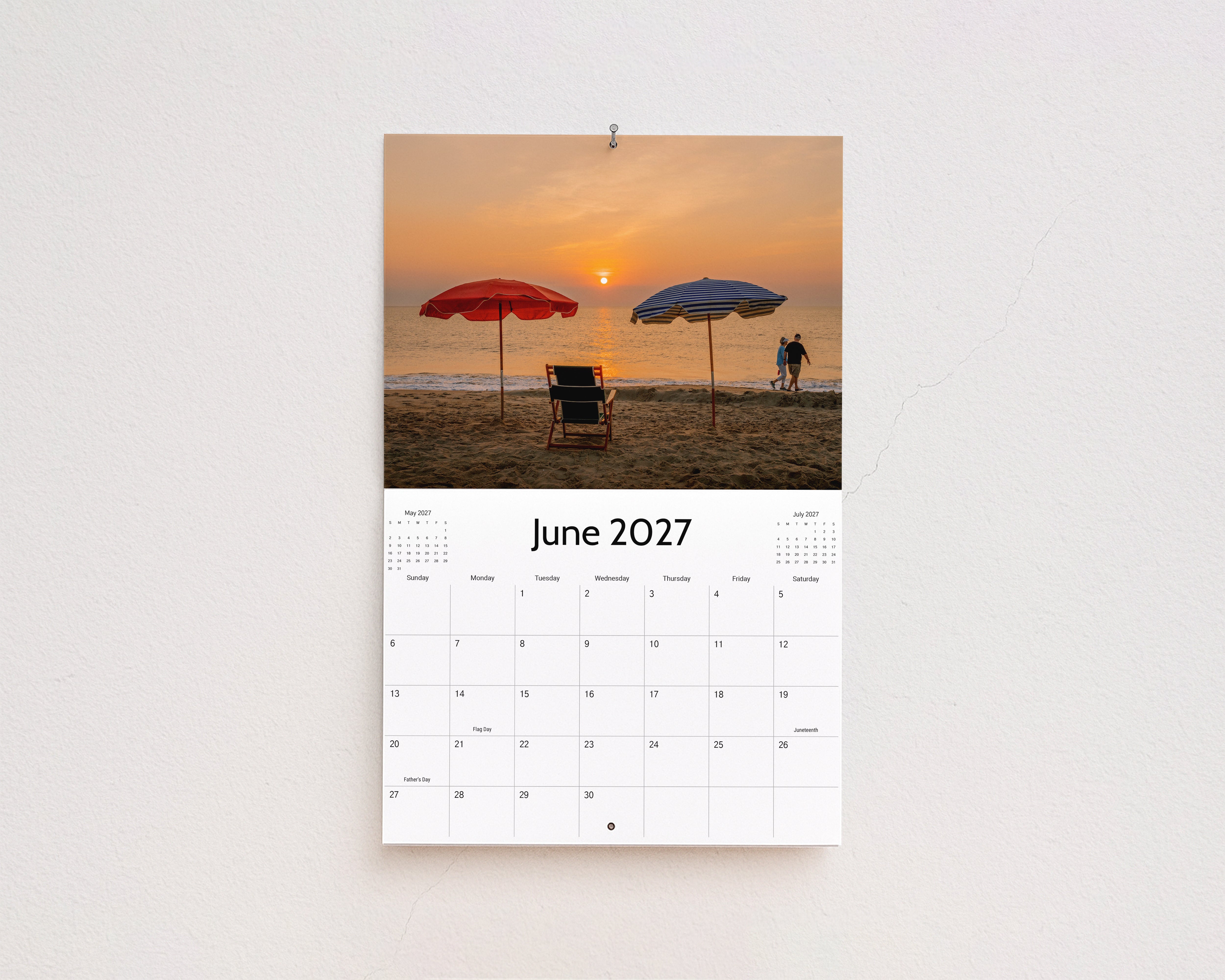 June 2027 page: red and blue beach umbrellas and a chair on the sand at sunrise