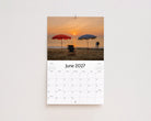 June 2027 page: red and blue beach umbrellas and a chair on the sand at sunrise